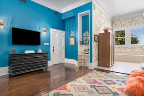 A room with a blue wall, a flat-screen TV mounted above a black console, a patterned rug, and a wooden cabinet. - The Loft on Main St, Balcony Views, Roman bathtub! (Fredericksburg)