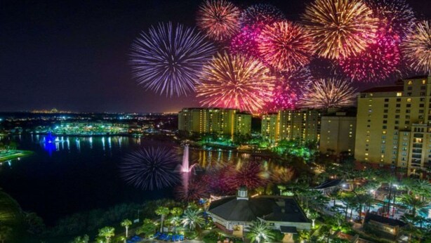 A resort with a swimming pool, palm trees, and a view of fireworks over a cityscape. - Great Value (Lake Buena Vista)