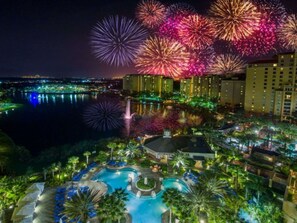 A resort with a swimming pool, palm trees, and a view of fireworks over a cityscape.