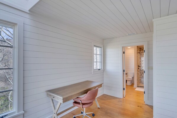 Une pièce aux murs recouverts de panneaux blancs, avec un bureau en bois, une chaise rose et une vue sur l’extérieur par une fenêtre.