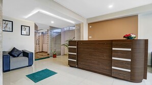 A modern reception area with a wooden reception desk, a grey sofa with a black cushion, a green rug, and a potted plant. - Treebo Elite Premium Homes (Bengaluru)