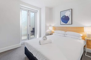 Une chambre d’hôtel avec un lit, une table de chevet, un tableau abstrait accroché au mur, une porte vitrée donnant sur un espace extérieur et une serviette sur le lit.