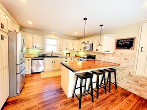 A kitchen with wooden flooring, white cabinets, a central island with stools, stainless steel appliances, and pendant lighting.