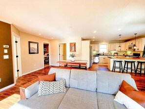 A modern living room with a grey sofa, wooden flooring, and a kitchen in the background.