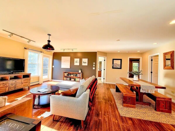 A spacious living room with a large flat-screen TV, a grey sofa, a wooden coffee table, and a dining area with a wooden bench and table.