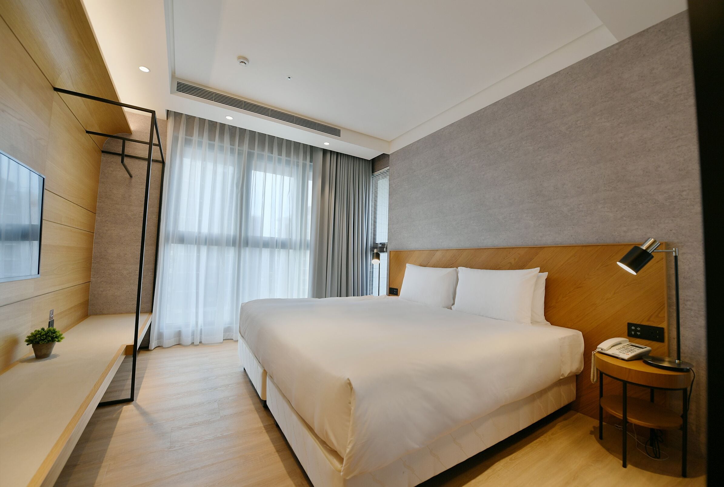 A modern hotel room with a large bed, a bedside table with a lamp, a small plant, and a window with curtains.