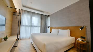 A modern hotel room with a large bed, a bedside table with a lamp, a small plant, and a window with curtains.
