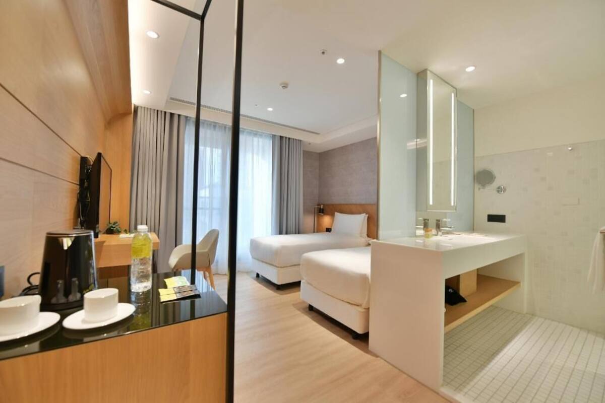 A hotel room with a bed, a desk with a coffee maker, a mirror, a bathroom with a shower, and a view of the city.