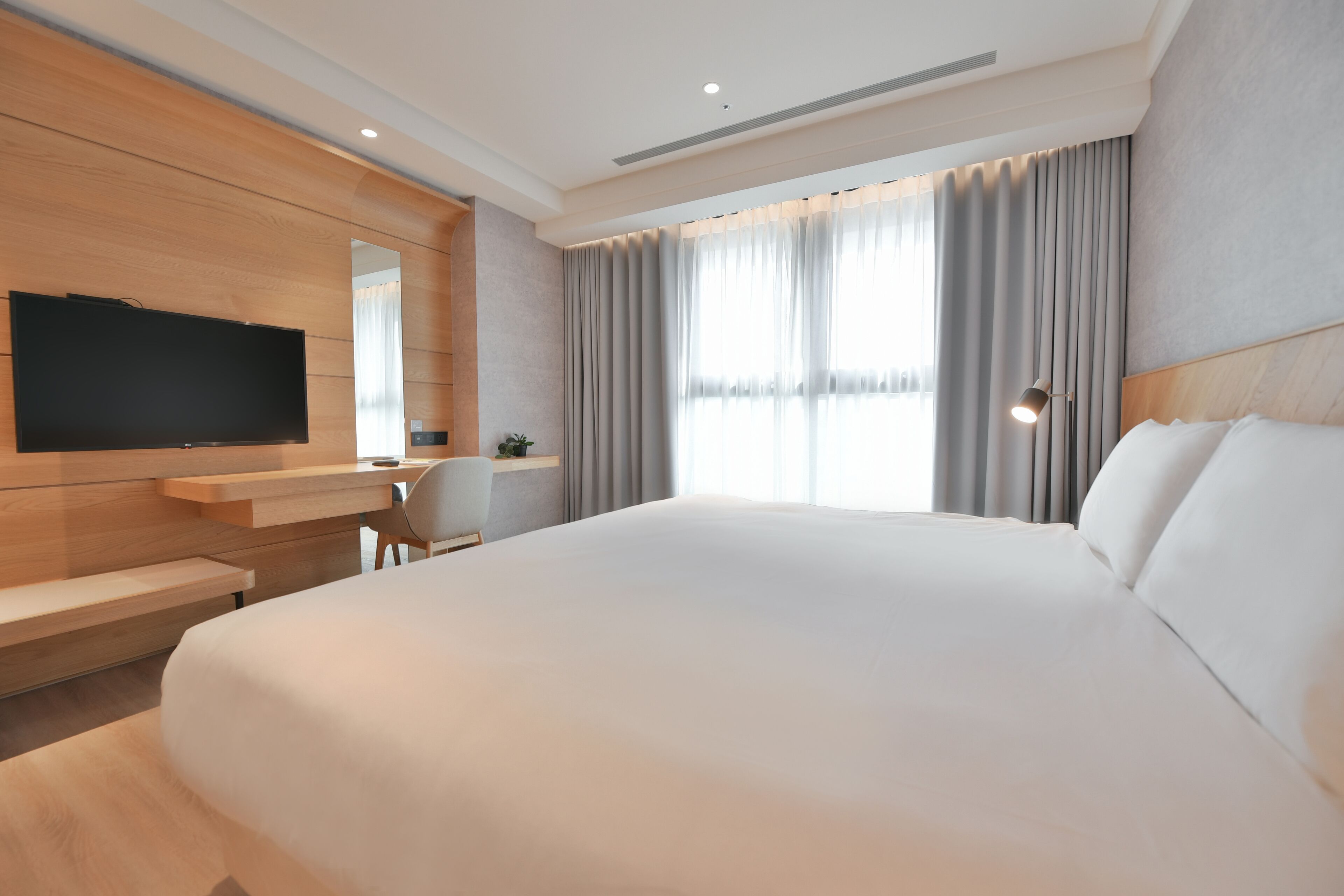 A modern hotel room with a large bed, a flat-screen TV mounted on the wall, a desk with a chair, and a large window with curtains.