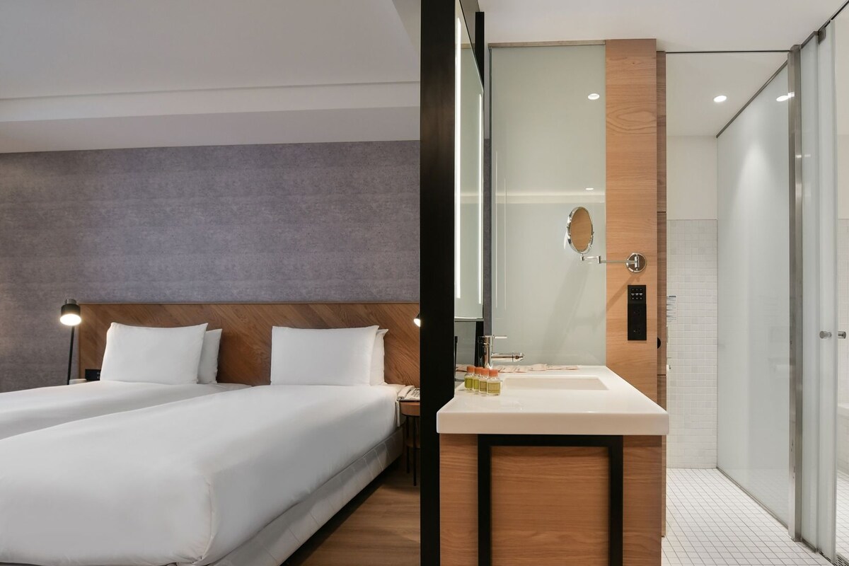 Une chambre d’hôtel moderne avec deux lits, une tête de lit en bois, un grand miroir et une salle de bain équipée d’un lavabo et d’une douche.