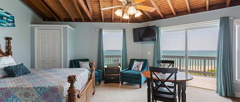 A bedroom with a bed, a desk, a chair, a TV, and a balcony with a view of the ocean.