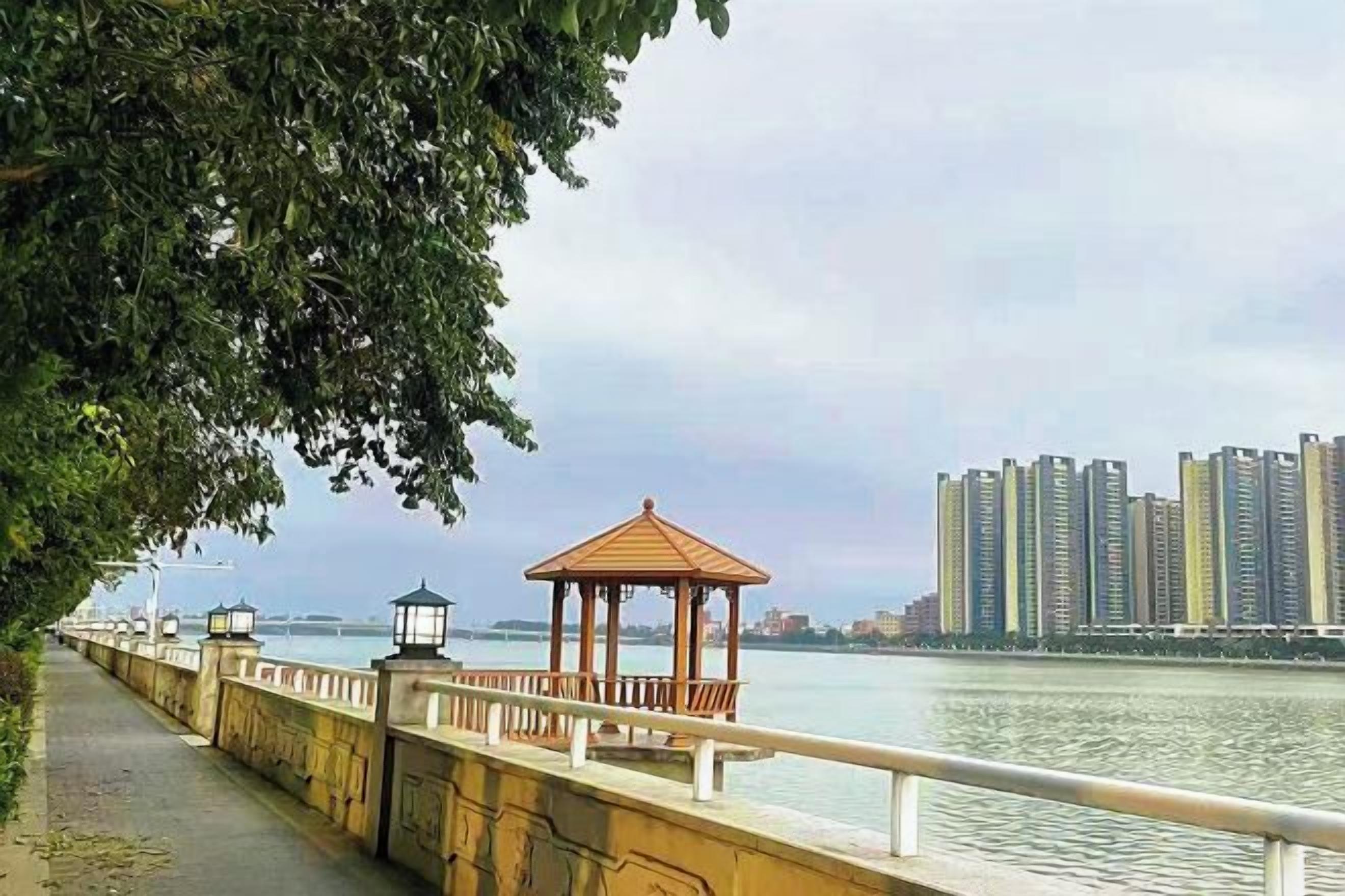 A riverside walkway with a gazebo and lamp posts, overlooking a body of water and high-rise buildings.