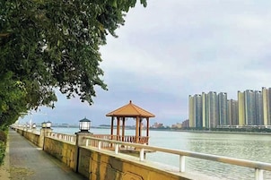 A riverside walkway with a gazebo and lamp posts, overlooking a body of water and high-rise buildings.