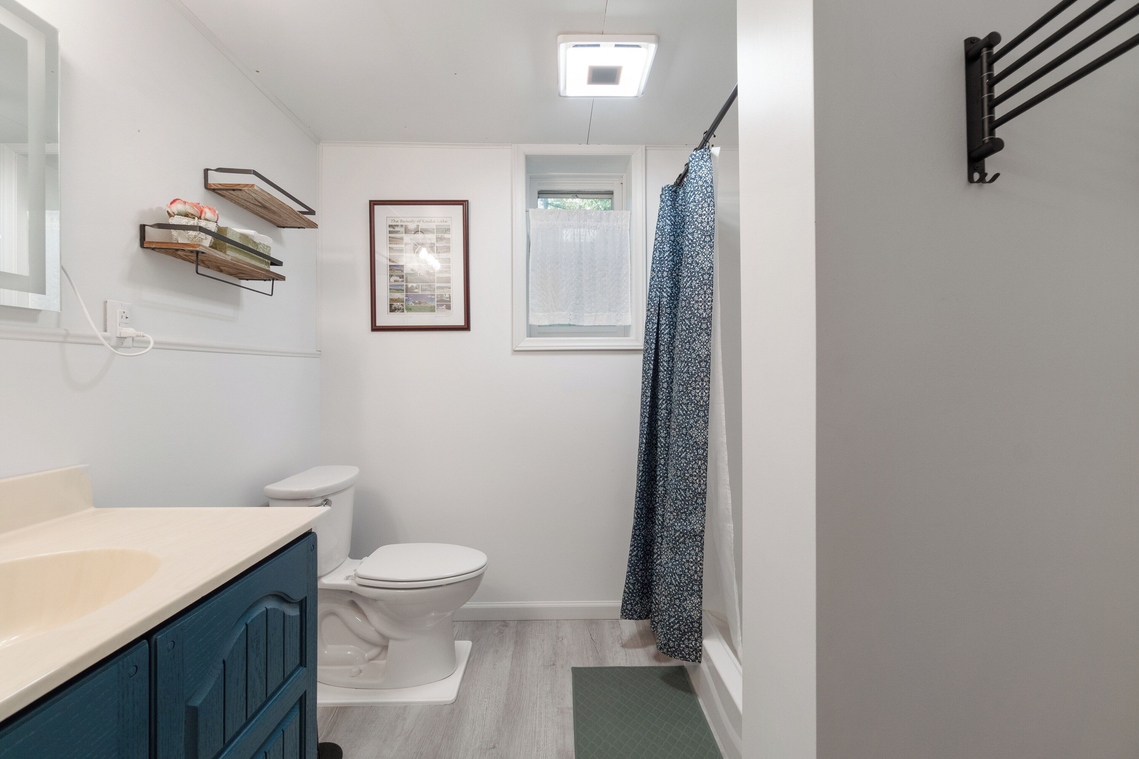 A bathroom with a white toilet, a sink with a beige countertop, a framed picture on the wall, a window with a white curtain, and a shower area with a patterned curtain.