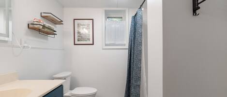 A bathroom with a white toilet, a sink with a beige countertop, a framed picture on the wall, a window with a white curtain, and a shower area with a patterned curtain.