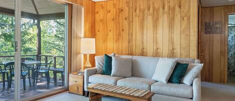 Une pièce avec un mur lambrissé en bois, un canapé blanc agrémenté de coussins turquoise, une table basse en bois et une porte en verre donnant sur une terrasse extérieure avec des chaises.