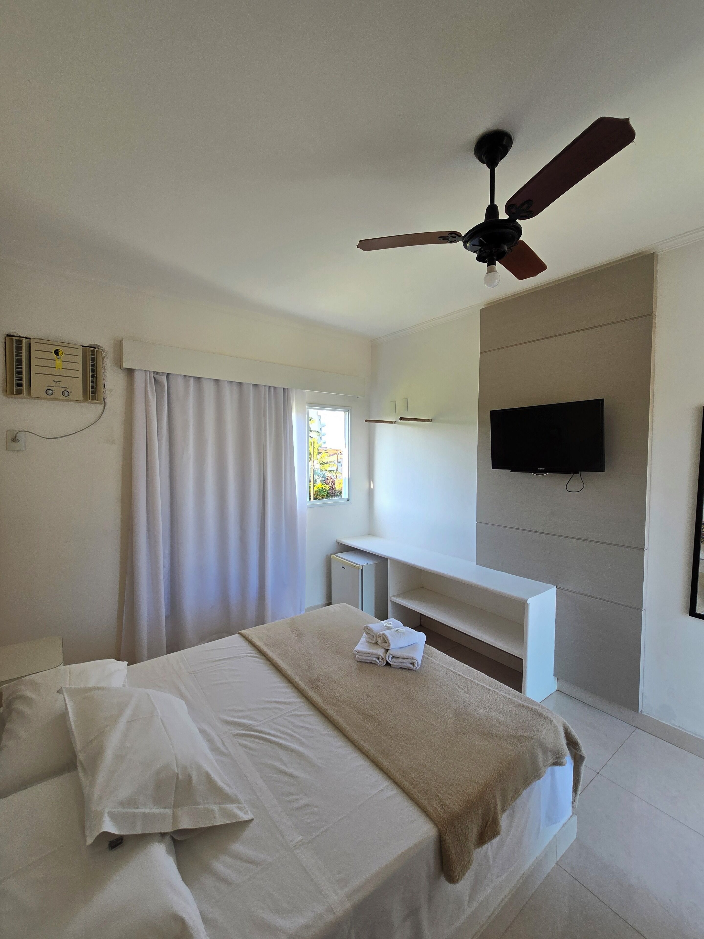 A hotel room with a bed, a ceiling fan, a TV mounted on the wall, and a window with curtains.