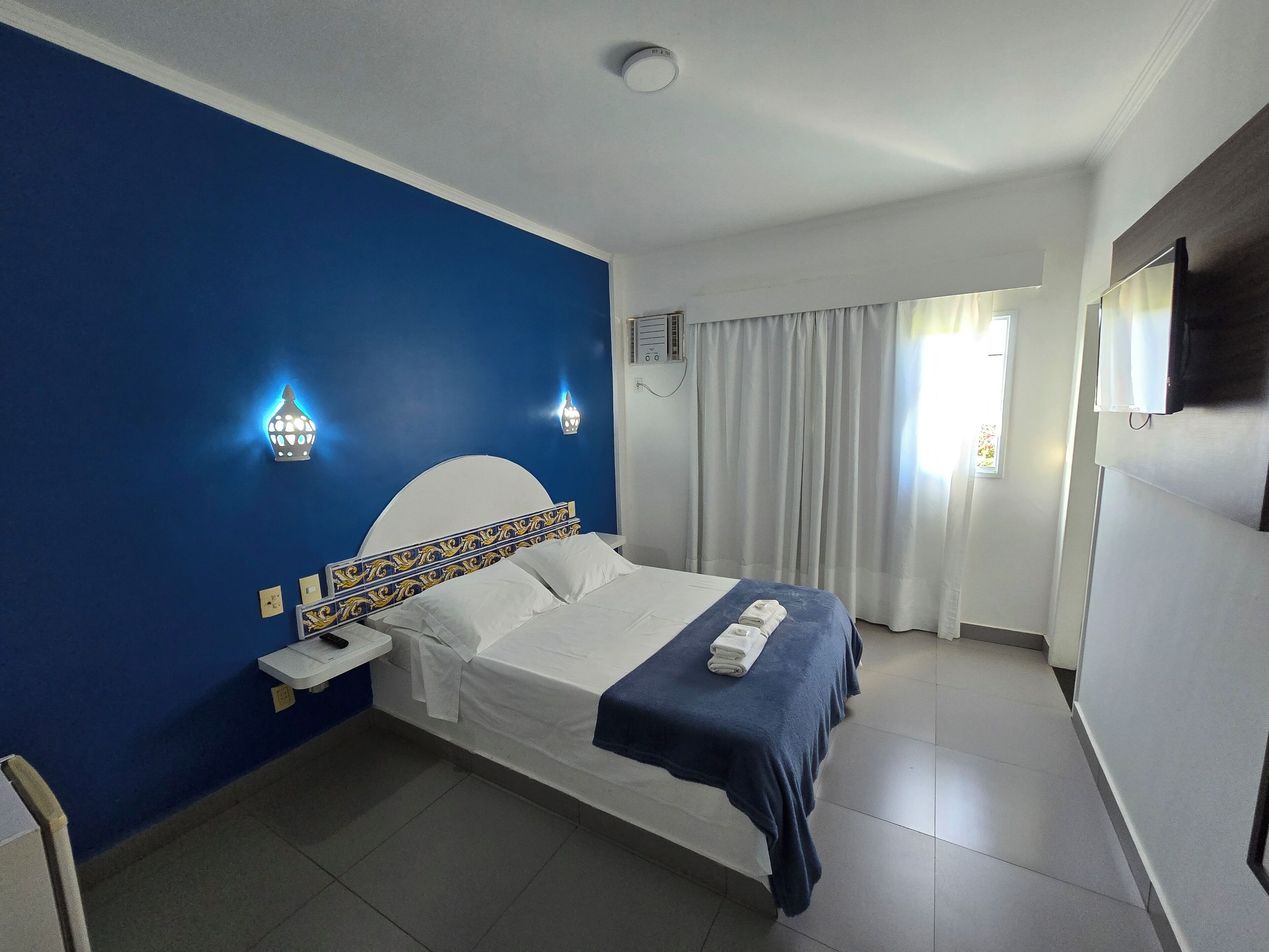Habitación de hotel con cama, mesita de noche, lámpara de pared, ventana con cortinas y televisor de pantalla plana.