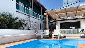 A swimming pool with a wooden deck, white chairs, and a covered patio area. - Pousada Elxadai (Guarapari)