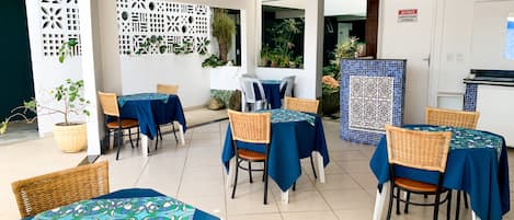Un área de comedor al aire libre con mesas cubiertas con manteles azules, sillas de mimbre y plantas en macetas.