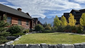 A log cabin-style building with a stone retaining wall, a well-maintained lawn, and a mountain backdrop. - L202 Canmore Vista- Free Parking, Hot tub, Pool, View, BBQ, 2BR, 2BT, Sleeps 6 (Canmore)