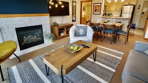 A modern living room with a fireplace, a dining area, and a kitchen. - L202 Canmore Vista- Free Parking, Hot tub, Pool, View, BBQ, 2BR, 2BT, Sleeps 6 (Canmore)