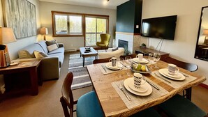 A modern living room with a dining area, a fireplace, and a TV mounted on the wall. - L202 Canmore Vista- Free Parking, Hot tub, Pool, View, BBQ, 2BR, 2BT, Sleeps 6 (Canmore)