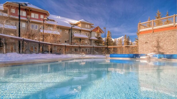 A clear pool with a wooden deck and a stone wall, surrounded by snow-covered buildings. - L202 Canmore Vista- Free Parking, Hot tub, Pool, View, BBQ, 2BR, 2BT, Sleeps 6 (Canmore)