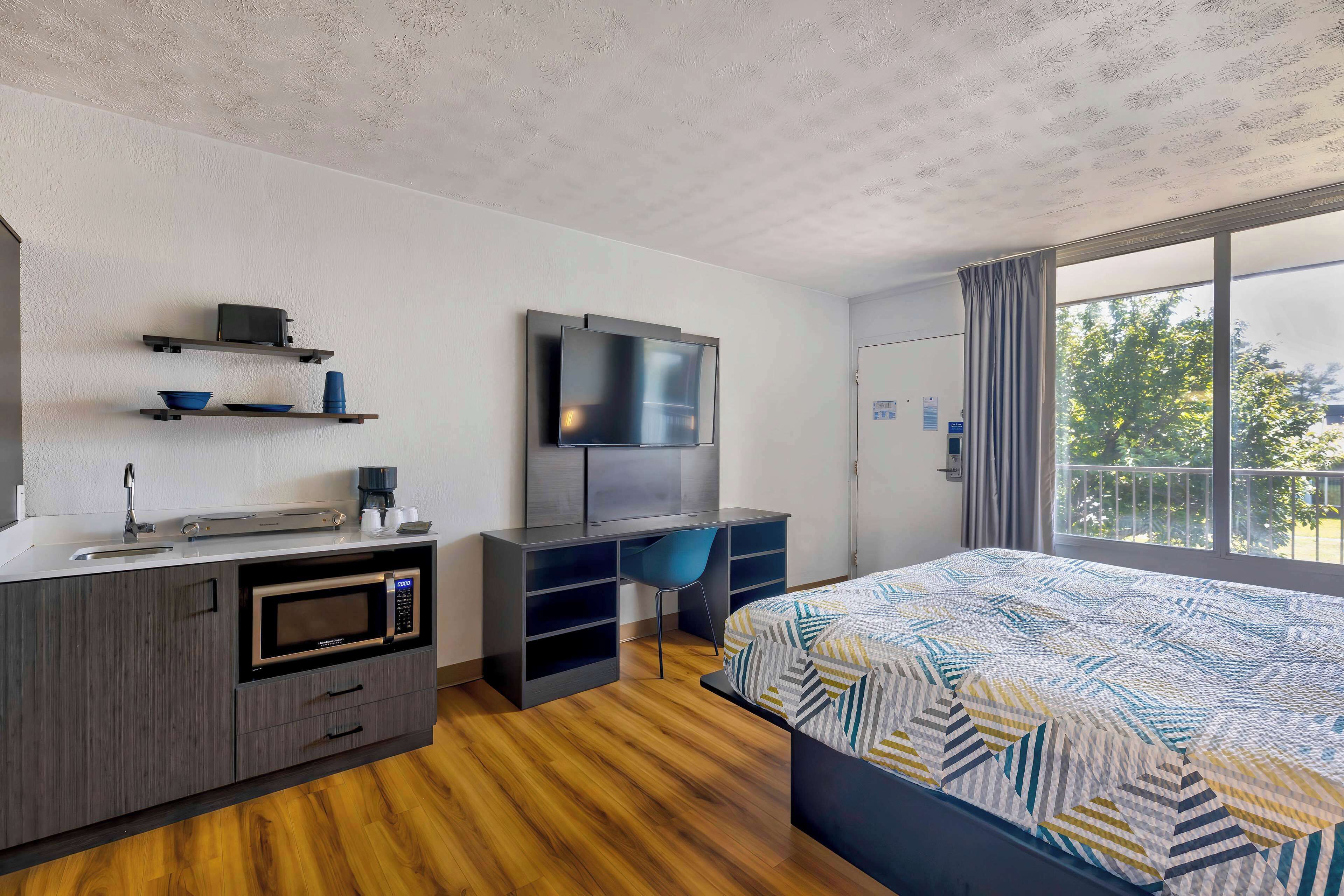 Ein modernes Hotelzimmer mit Bett, Schreibtisch, Flachbildfernseher, Mikrowelle, Kaffeemaschine und Fenster mit Blick auf Bäume.