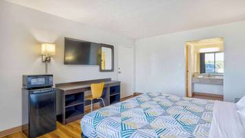 Habitación de hotel con cama, escritorio con silla, refrigerador pequeño, microondas, televisor montado en la pared y ventana con vista.