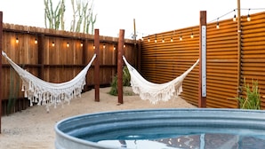 Un giardino sul retro con recinto in legno, due amache, una vasca idromassaggio in metallo e luci decorative appese.