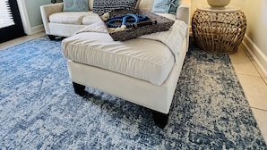 A living room with a beige sofa, a blue patterned rug, a wicker side table with a lamp, and a painting of a beach scene. -  Bayside Cottage 4 miles to Destin, Shopping & Beaches, minutes to Elgin AFB (Niceville)
