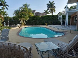 A swimming pool with lounge chairs and a table, surrounded by palm trees and a hedge.