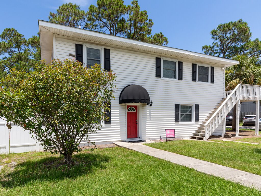 A two-story white house with a red door, a small porch, and a staircase leading to a covered area.
