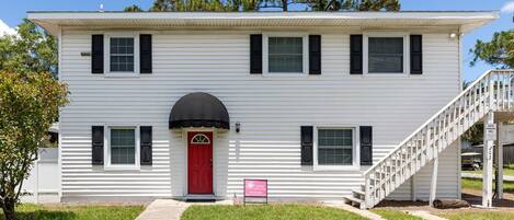 A two-story white building with a red door, black shutters, and a staircase leading to the second floor.