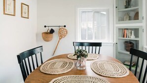 A dining area with a round wooden table, black chairs, woven placemats, a hanging basket, and a window with blinds.