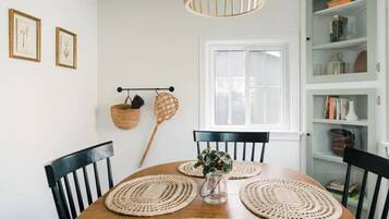 Un coin repas avec une table ronde en bois, des chaises noires, des sets de table en fibres tressées, un panier suspendu et une fenêtre avec des stores.