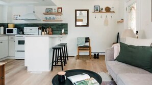 A modern kitchen with white cabinets, a central island, and a dining area with stools. There is a sofa and a lamp in the living area.