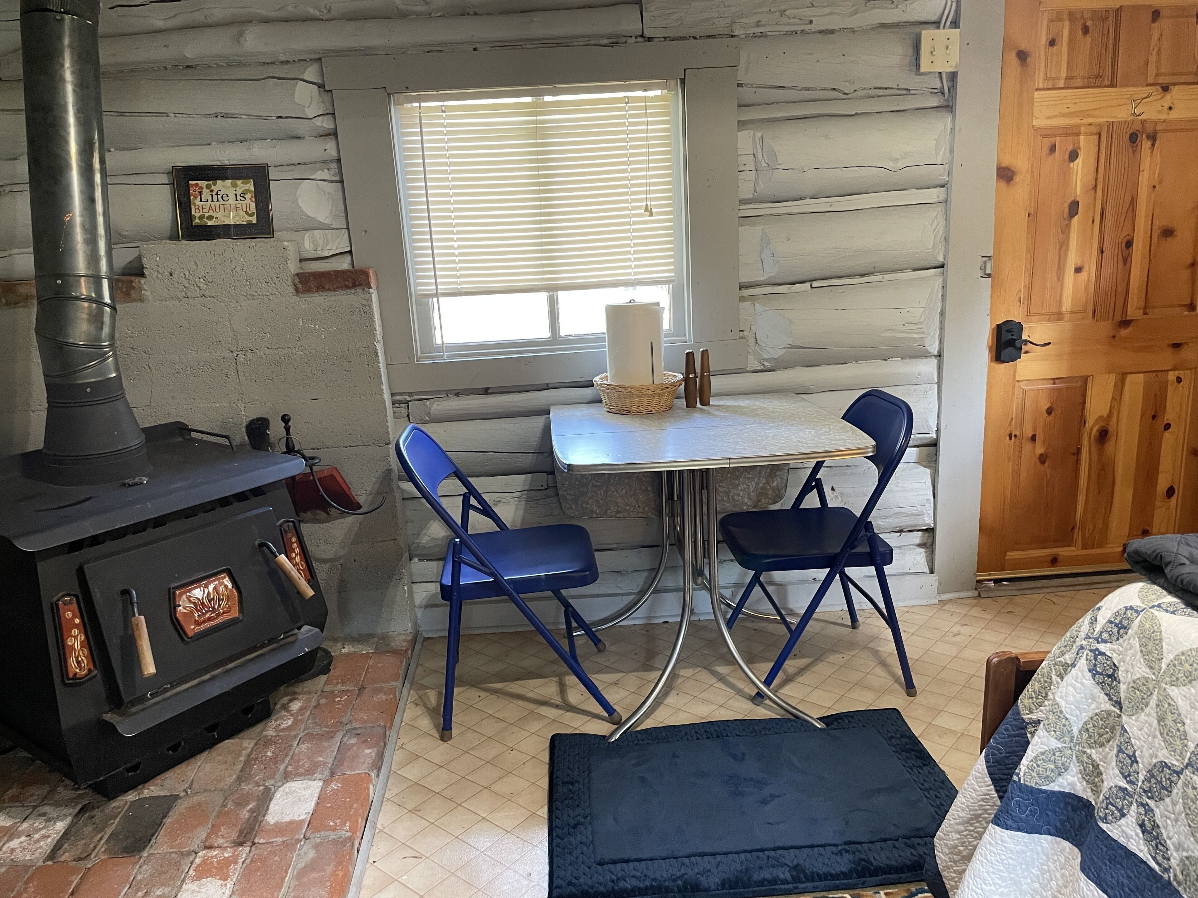 L’intérieur chaleureux d’un chalet avec un poêle à bois, une table à manger avec des chaises, une fenêtre avec des stores et une porte en bois.
