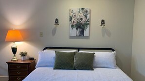 A neatly made bed with a white duvet and two green pillows, a bedside lamp, a small plant, and a painting of flowers on the wall.