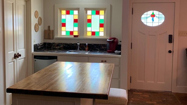 A kitchen with a wooden island, two windows with colorful stained glass, a white door with a circular window, and a stone floor.