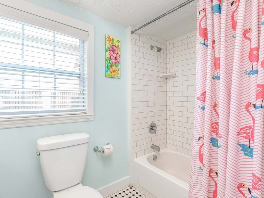 A bathroom with a toilet, a shower with white tiles, and a window with blinds.