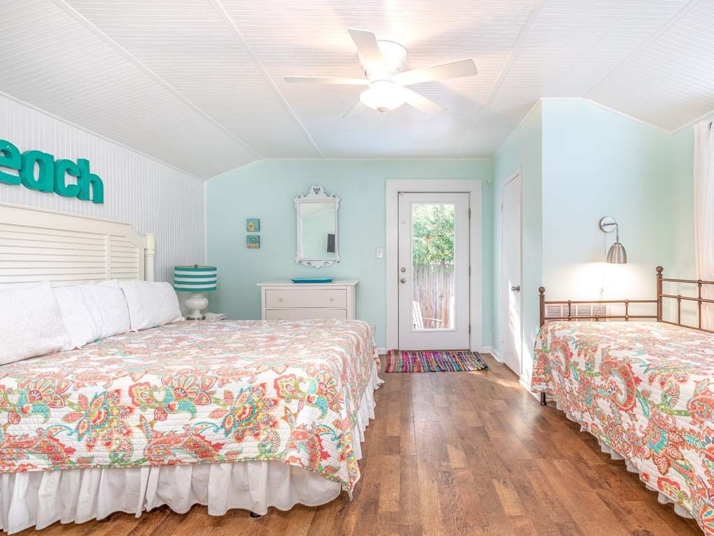 A bedroom with two beds, a ceiling fan, a door with a view, and a small table with a lamp.