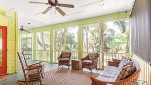 A covered porch with wicker furniture, a ceiling fan, and large windows overlooking a wooded area. - Blue Skies by Tybee Vacation Rentals (Tybee Island)