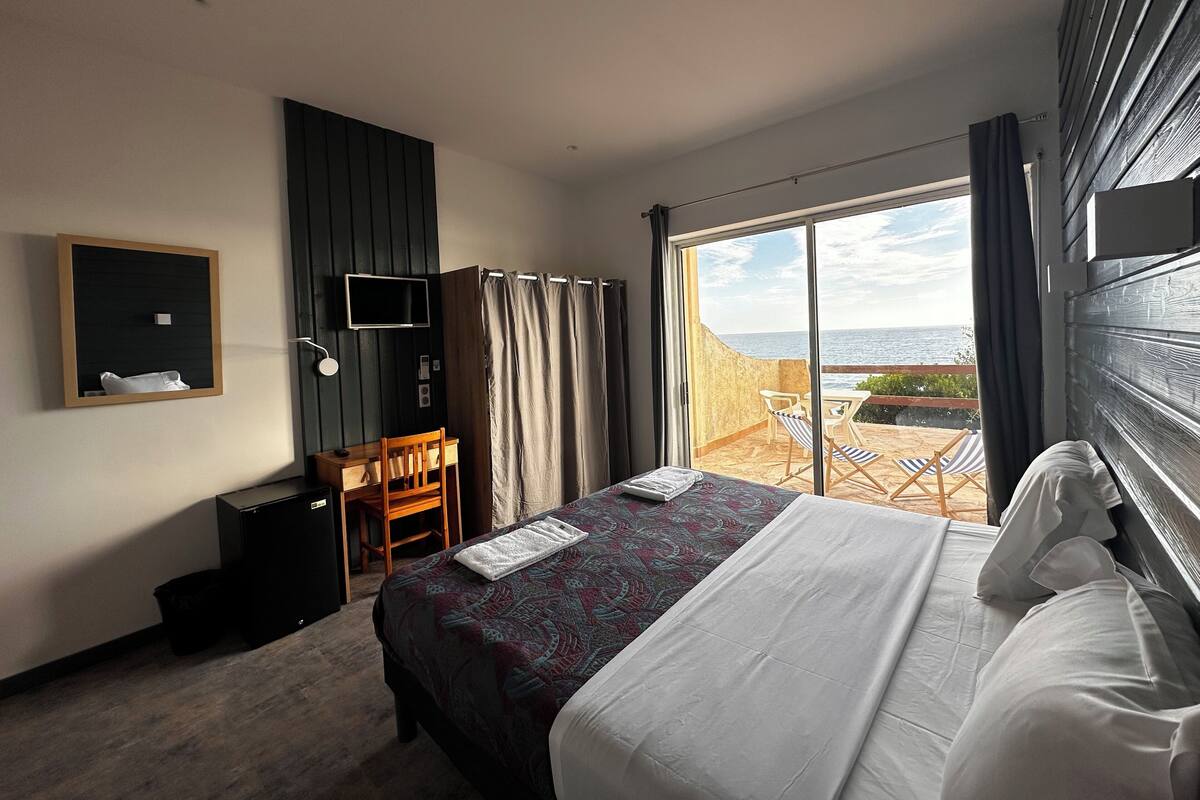 A hotel room with a bed, a desk, a chair, a TV, and a balcony with a view of the sea.