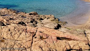Paisaje costero con terreno rocoso en primer plano, una playa arenosa y una colina con vegetación al fondo.