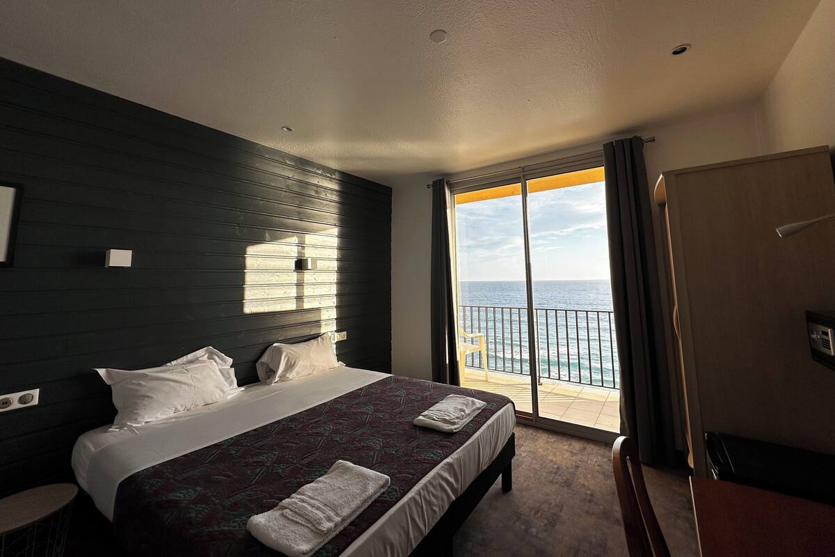 A hotel room with a bed, a small table, a chair, a balcony with a view of the sea, and a sliding glass door.