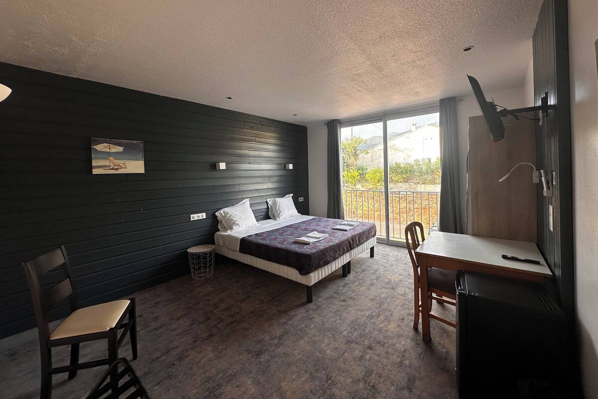 A bedroom with a bed, a desk, a chair, a small table, and a balcony with a view.