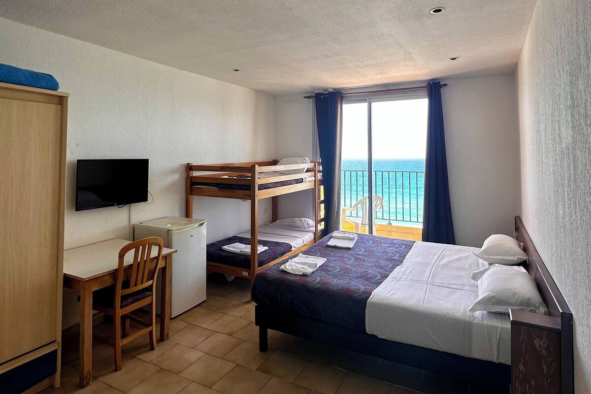 A compact hotel room with bunk beds, a desk, a chair, a television, and a balcony with a view of the sea.