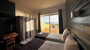 Habitación de hotel con dos camas, un escritorio y un televisor. Hay una puerta corrediza de vidrio que da a un espacio al aire libre con vista al mar.